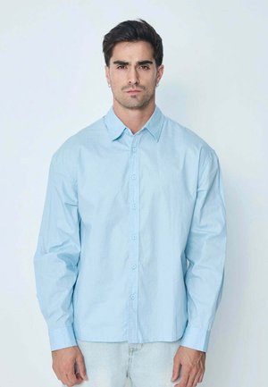 Homme aux cheveux foncés portant une chemise bleue claire à boutons et un jean clair debout devant un fond blanc uni.