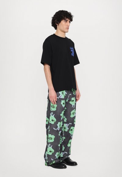 Schwarzes T-Shirt mit blauer Grafik, kombiniert mit grauen Hosen mit grünen Blumenmustern. Das Modell trägt schwarze Schuhe und steht vor einem weißen Hintergrund.