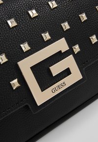 Sort tekstureret syntetisk håndbag med guldtonede firkantede nitter og et fremtrædende metallisk logo. 