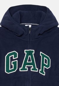 Felpa con cappuccio in pile blu navy, dotata di alto collo, zip completa e grande logo "GAP" ricamato in bianco con dettagli verdi. Texture morbida.