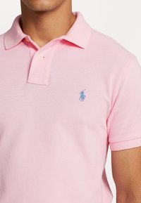 Rosa polo skjorta i strukturerad tyg med tre knappar i placket och en broderad blå logotyp på vänster bröst.