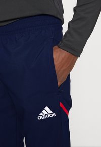 Siniset urheiluhousut, jotka on valmistettu kevyestä kankaasta ja joissa on joustava vyötärönauha, sivutaskut sekä valkoinen Adidas-logo punaisilla yksityiskohdilla.