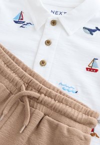 Weiße Baumwoll-Poloshirt mit bestickten Booten und Wellen, dazu braune Shorts mit Kordelzug aus strukturiertem Stoff. Die Knöpfe sind goldfarben.