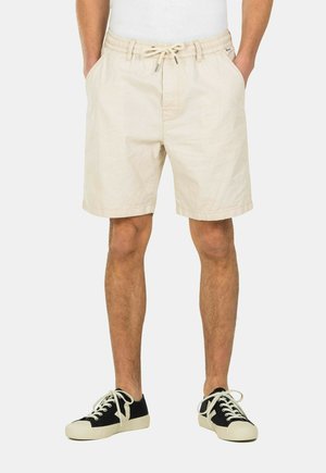 Beige cotton shorts with an elastic waistband and drawstring. Side pockets and a relaxed fit. Paired with black sneakers and white laces.