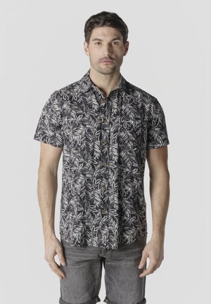 Hombre de pie mirando hacia adelante, vestido con una camisa negra de manga corta con botones y estampado de hojas blancas, y pantalones cortos de color gris oscuro.