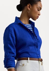 Camisa de linho azul com frente abotoada e mangas arregaçadas. Combinada com uma blusa às riscas e calças brancas, acentuada por um cinto castanho.