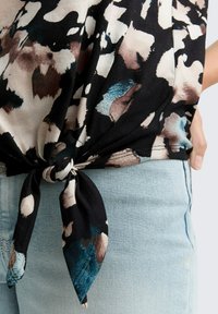 Top floreale nero, beige e blu legato in un nodo in vita sopra jeans in denim blu chiaro, con una mano appoggiata sui fianchi.
