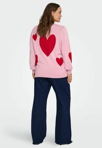 Světlá růžová svetr s červenými vzory srdcí na zadní straně, dlouhými rukávy a volným střihem. Kombinován s tmavě modrými širokými džíny.