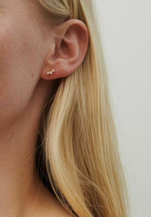 Lähikuva naisen korvasta, jossa on kolme pientä helmiruusuketta ja pitkä suora vaalea tukka yksinkertaista vaaleaa taustaa vasten.