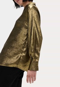 Camisa de manga larga metálica dorada con puños abotonados y cuello, con un tejido texturizado y brillante que refleja la luz.