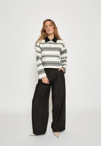 Prúhovaná polokošeľa s dlhým rukávom v bielej, čiernej a sivej farbe s čiernym golierom, skombinovaná s voľnými čiernymi nohavicami a špicatými béžovými topánkami.