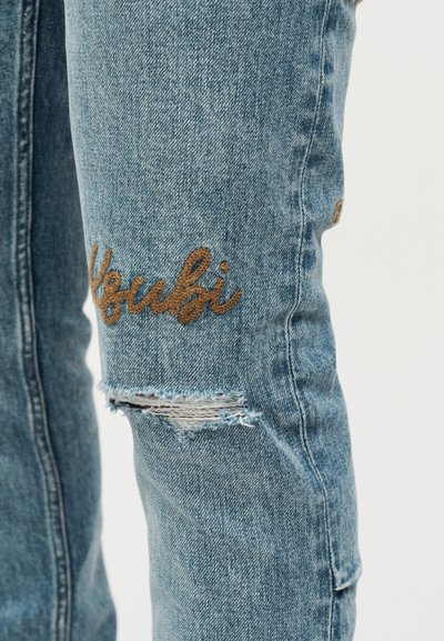 Κοντινό πλάνο μπλε τζιν παντελονιού με σκισμένο γόνατο και καφέ κεντημένο κείμενο "Youbi" στο μηρό.