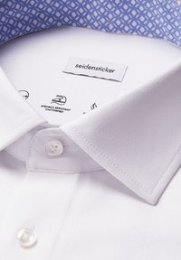 Colletto della camicia bianca con bottone chiuso, piegato in modo ordinato, che mostra l'etichetta del marchio "seidensticker" e l'icona per le istruzioni di stiratura.
