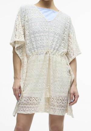 Kobieta ubrana w kremową, szydełkowaną koronkową sukienkę z krótkimi rękawami i talią ze sznurkiem, na jasnoniebieskim topie.