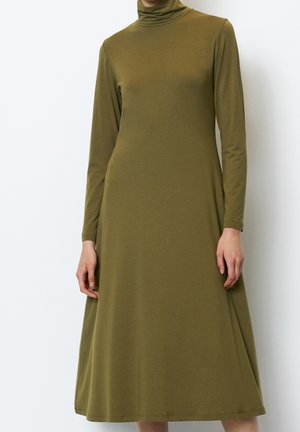 Woman wearing an olive green long-sleeve turtleneck midi dress standing against a white background.