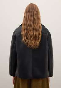 Black leather jacket with a relaxed fit, visible seams, and a wide collar. Hair is long and wavy, and the bottom half is covered by a yellow skirt.