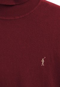 Vinröd tröja i stickat med ribbad polokrage och en liten beige broderad logotyp av en golfspelare. Mjuk textur och figursydd design.