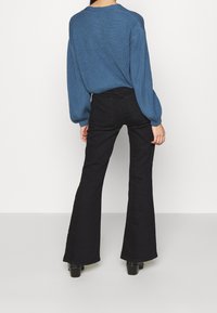 Blauwe geribde trui met brede mouwen, gecombineerd met zwarte flared jeans en chunky haklaarzen. Neutrale achtergrond benadrukt de outfit.