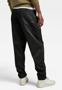 Pantalons noirs en tissu lisse, avec une coupe décontractée, des poches arrière et une étiquette en patch. Associés à des sneakers blanches.