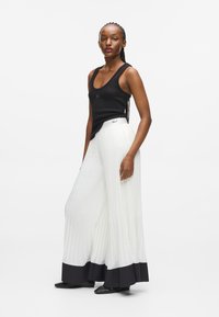 Czarny top na ramiączkach w zestawieniu z białymi szerokimi spodniami z plisami, które mają czarny dolny brzeg. Gładki materiał z subtelną fakturą; minimalistyczne akcenty w designie.