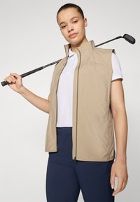 Beige sleeveless vest in lightweight fabric, featuring a high collar, zip closure, and side pockets, worn over a white polo shirt and blue pants.