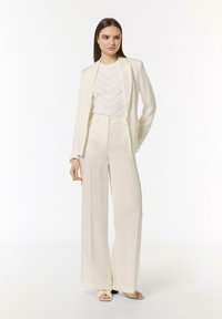 Cremfarbener Blazer und weiter Hosen aus einem leichten Stoff, kombiniert mit einem strukturierten weißen Oberteil mit einem Zickzackmuster und beigen Sandalen.