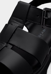 Μαύρο σανδάλι με φαρδιές λουρίδες σε ματ φινίρισμα. Περιλαμβάνει μεταλλική πόρπη και υφασμένο πέλμα. Έμφαση σε στοιχεία μοντέρνου σχεδιασμού.