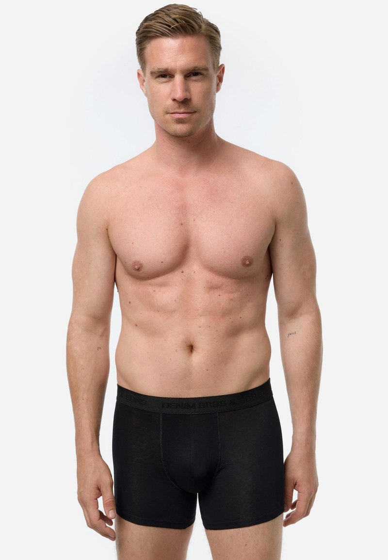 Bóxers negros hechos de material suave con una cinturilla elástica que presenta un logo. Modelo de pie, sin camiseta, con un fondo neutro.
