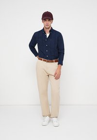 Férfi burgundi sapkában, tengerészkék csíkos ingben, bézs nadrágban, barna övvel és fehér tornacipőben áll a fehér háttér előtt.