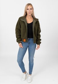 Oliwkowa polarowa kurtka z zamkiem z przodu, ściągaczami przy mankietach i kołnierzem z panelu. Połączona z niebieskimi skinny jeansami i białymi sneakersami.