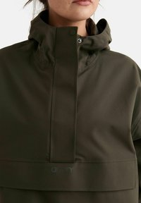 Gros plan sur une personne portant une veste à capuche vert olive avec un col montant et un rabat boutonné couvrant la fermeture éclair.