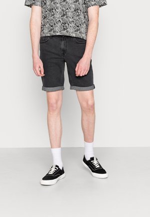 Person iført sorte opkørte denimshorts, sorte og hvide sneakers, hvide sokker og en mønstret kortærmet skjorte, der står på en grå gulv.