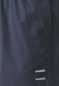 Marineblå shorts laget av lett stoff, med en jevn tekstur og to hvite "SAMSØE" logoer nær kanten.