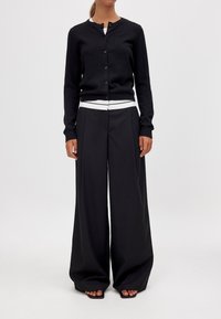 Schwarze Strickjacke mit rundem Ausschnitt und Knöpfen, kombiniert mit weiten schwarzen Hosen und einem Top mit weißen Streifen darunter.