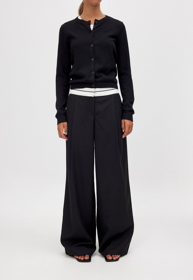 Schwarze Strickjacke mit rundem Ausschnitt und Knöpfen, kombiniert mit weiten schwarzen Hosen und einem Top mit weißen Streifen darunter.