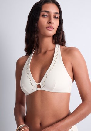 Femme portant un haut de bikini blanc style halter avec de petits clous dorés le long des bords, debout devant un fond clair uni.