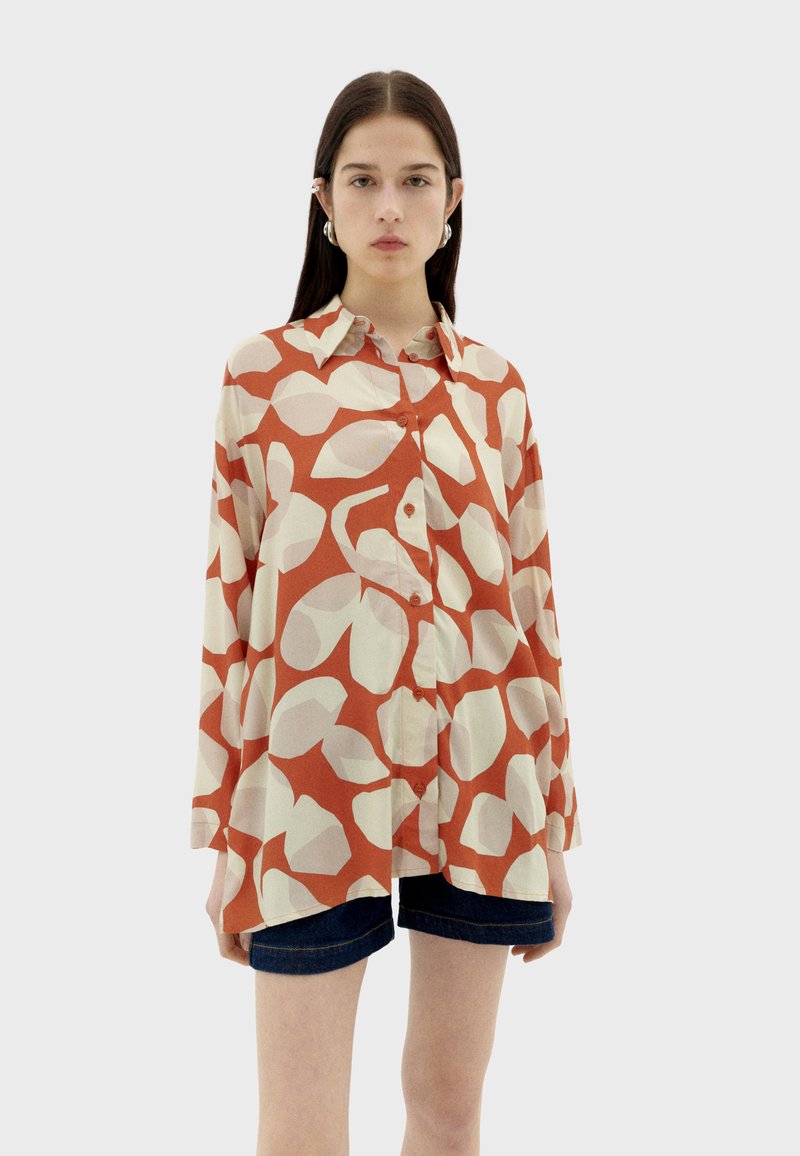 Lange mouwen shirt in crème met groot roestkleurig abstract patroon, knoopsluiting aan de voorkant en klassieke kraag. Gecombineerd met denim shorts.