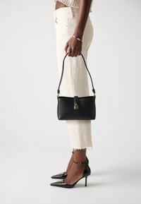 Sac en cuir noir avec un accent de serrure ton or, port�é par une personne en pantalon court crème et des escarpins noirs à bout pointu.