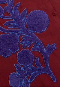 Rode stof met complexe paarse bloemenbroderie, voorzien van gestructureerde lussen en blauwe kralenaccenten, wat een gedetailleerd, gelaagd ontwerp creëert.