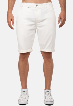 Mann trägt hochgekrempelte off-white Shorts und weiße niedrige Canvas-Sneaker, steht mit entspannten Armen.