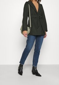 Tmavě zelený kabát s kapucí a zavazovacím pásem, vrstvený přes béžový svetr, doplněný modrými džínami a černými kotníkovými botami na podpatku.