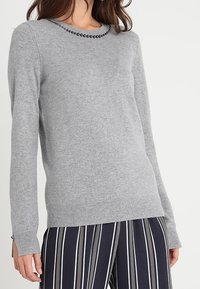 Pull gris en maille douce avec manches longues, encolure ronde ornée de perles noires, ourlet ajusté, associé à un pantalon rayé.