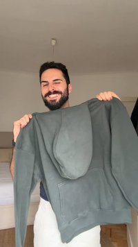 Homme souriant aux cheveux foncés et à la barbe, tenant un sweat à capuche gris dans une chambre lumineuse et minimaliste.
