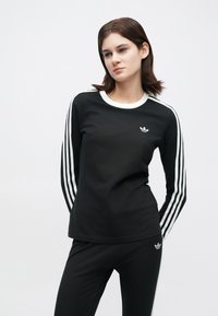 Žena v čiernom adidas teplákovom Oblečení s bielymi pruhmi na rukávoch a nohaviciach, stojí s rukami za chrbtom na jednoliatom pozadí.