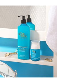 Bouteilles de lait autobronzant Blue Bondi Sands et lotion pour le visage sur une étagère blanche, avec des boucles d'oreilles dorées et un recourbe-cils à proximité.
