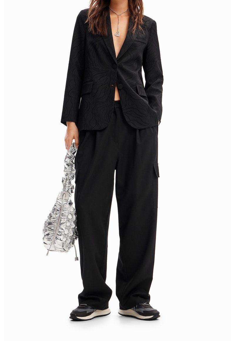 Femme portant un blazer noir à motifs, un pantalon noir ample, des baskets noires, un collier argenté et tenant un sac à main argenté brillant froncé.