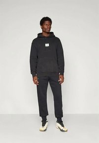 Muž v černé mikině s kapucí, černých teplákových kalhotách a bílých teniskách stojící na jednobarevném světlém pozadí.