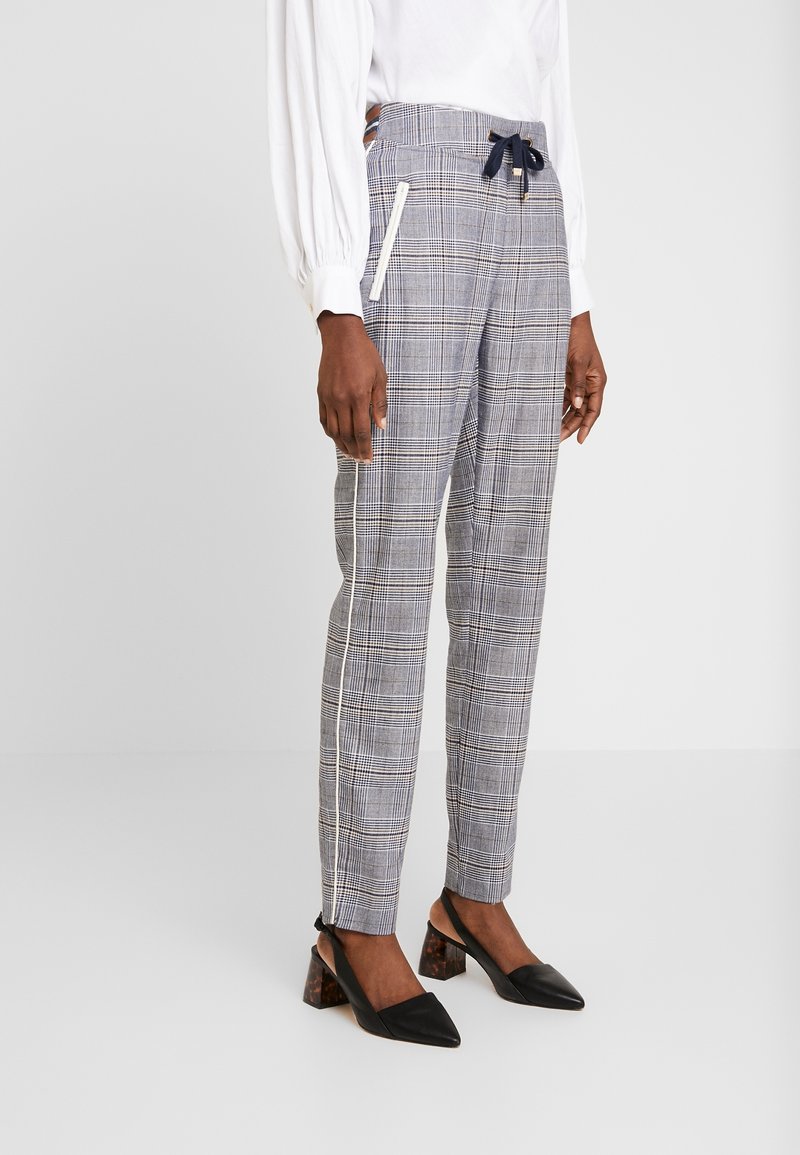 Pantalon slim à carreaux gris avec passepoil blanc sur les côtés et poches zippées, porté avec des escarpins slingback noirs et un chemisier blanc à manches longues.