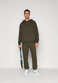 Σετ με παλαιολιθικό πράσινο φούτερ με κουκούλα και φόρμες, με άνετη εφαρμογή, ρυθμιζόμενη κουκούλα με κορδόνι και λεία υφή. Κρατάει ρακέτα του τένις.