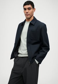 Ναυτικό φλιτζάνι με φερμουάρ και μία τσέπη στο στήθος, τοποθετημένο πάνω από ένα ανοιχτό πράσινο πουλόβερ, σε συνδυασμό με γκρι παν trousers, με χαλαρή εφαρμογή.